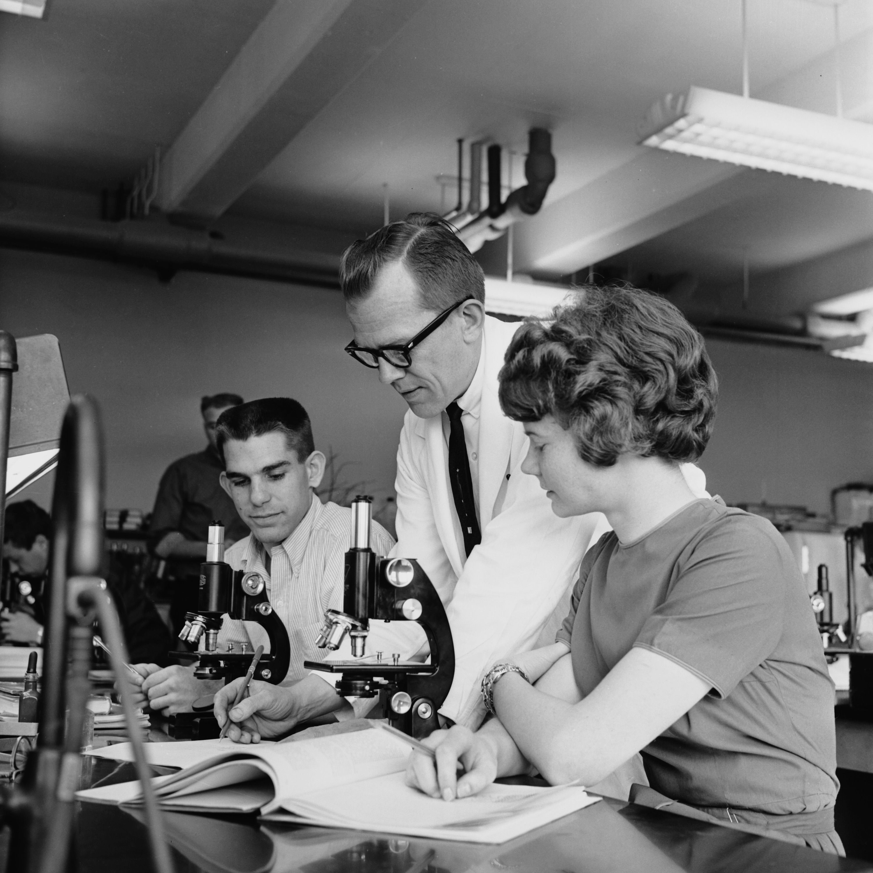 A black and white photo of two students at microscopes and a professor teaching them.