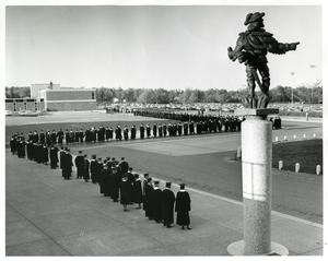 A black and white photo of students lined up in Ordean Court next to Sieur Du Luth, wearing graduation regalia.