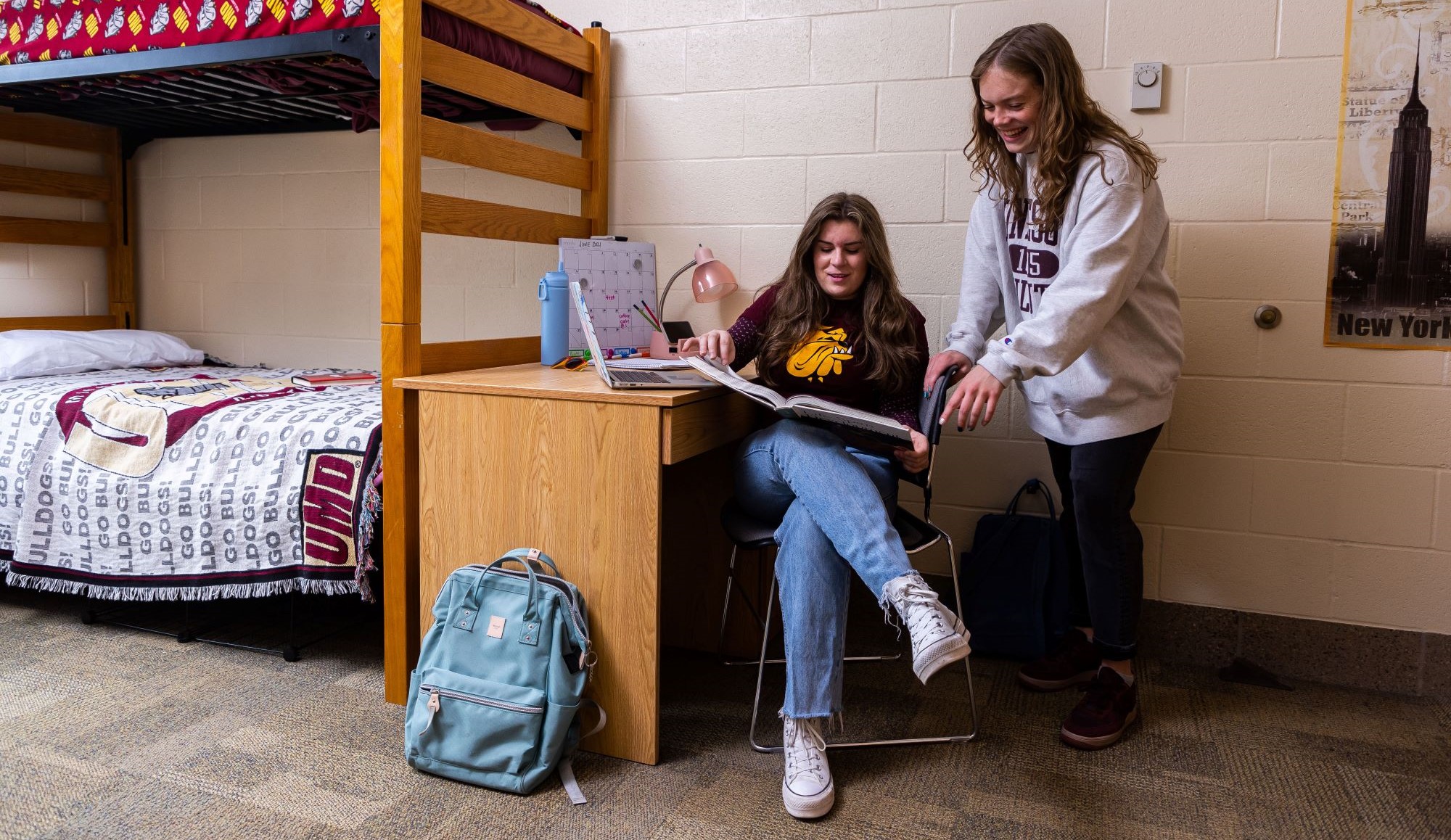 Two students studying in a dorm room with a textbook and a laptop