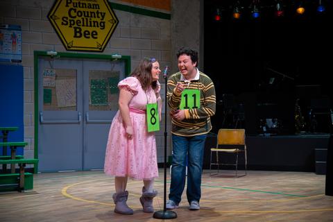 Two people on a stage, wearing numbers, with a sign in the background reading "Putnam County Spelling Bee."