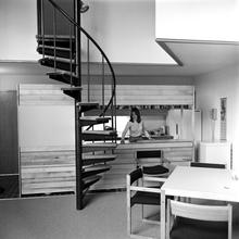 A Black and white photo of a young woman in a studio apartment, looking through an open kitchen, with a spiral staircase visible.
